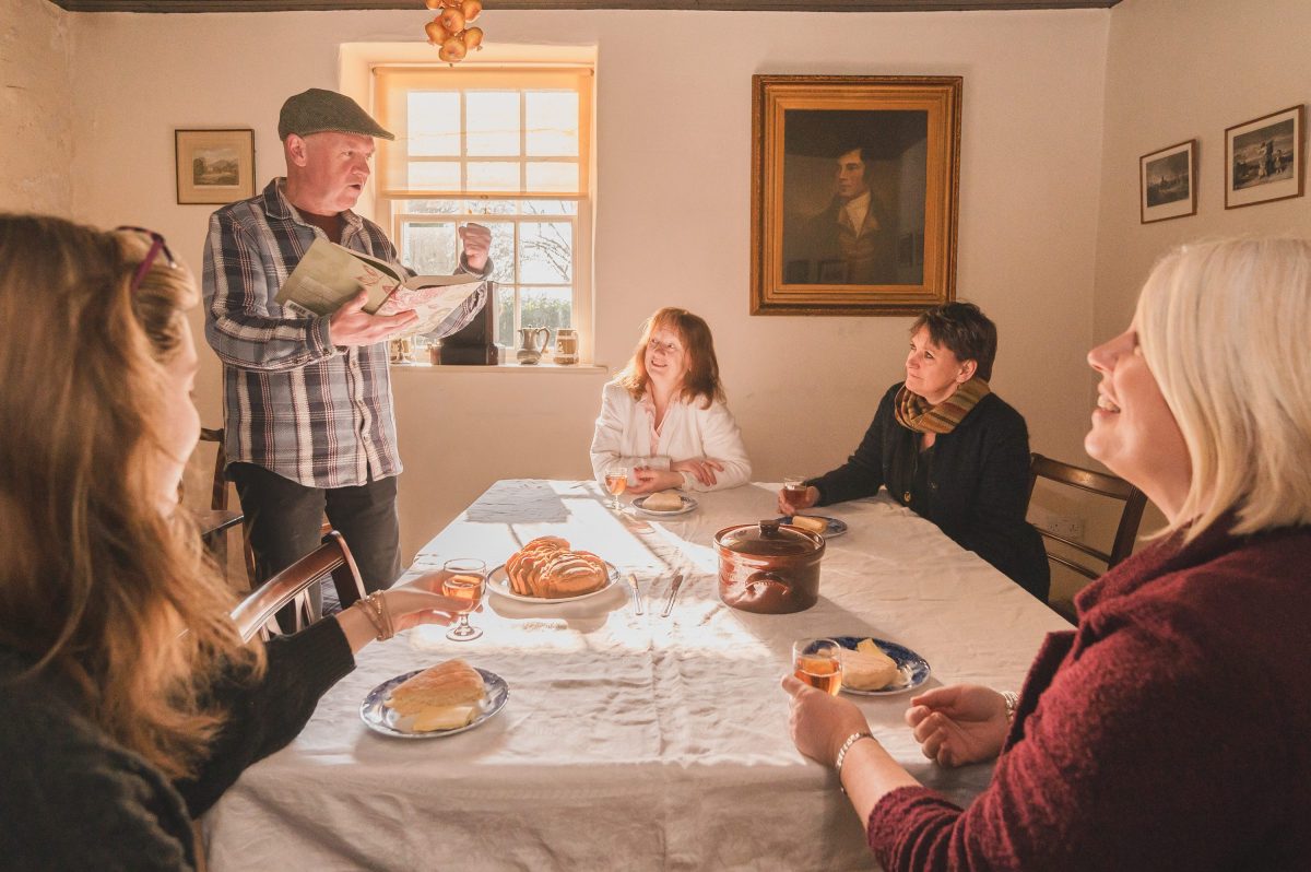 A man telling stories of Robert Burns to 4 visitors as they eat Bannocks and drink drams around a table.