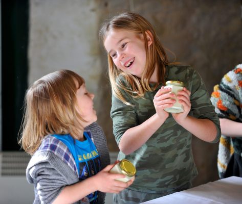 2 young girls smiling at each other whilst making butter in a jar